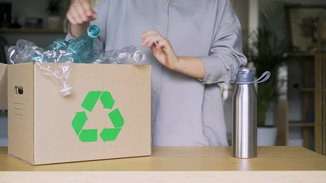 woman blogger in grey shirt puts empty plastic bottles into cardboard box with recycling sign and shows thumb-up near metal thermos at table closeup. Zero waste concept, reusable things