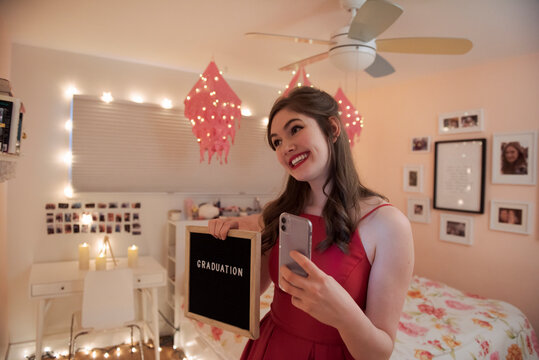 Happy teenage girl taking selfie with graduation sign in bedroom