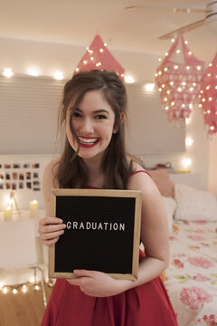 Portrait of smiling teenage girl holding graduation sign in bedroom
