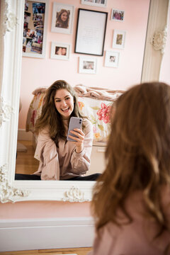 Happy teenage girl with smart phone taking selfie at bedroom mirror