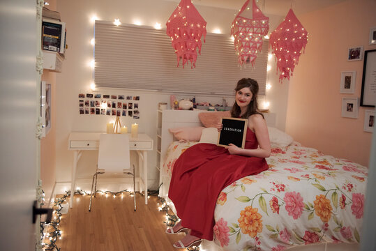 Portrait teenage girl in red dress holding graduation sign on bed
