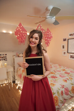 Portrait happy teenage girl holding graduation sign in bedroom