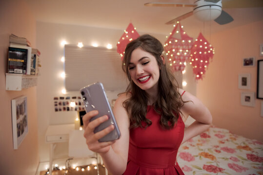 Happy teenage girl in red graduation dress video chatting in bedroom