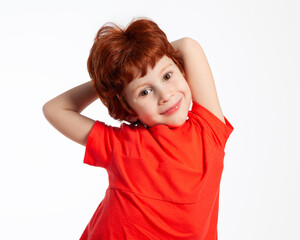 A beautiful European red-haired boy in a red t-shirt on a white background. The child smiles, performs a physical exercise-leaning to the sides. Physical education, fitness, sports.