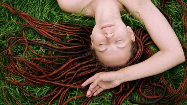 girl with red dreadlocks lying on the green grass, squinting from the sun, smiling. view from above.