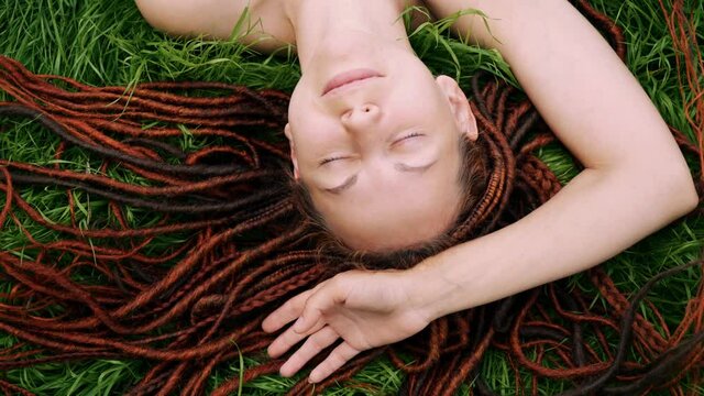 girl with red dreadlocks lying on the green grass, squinting from the sun, smiling. view from above.