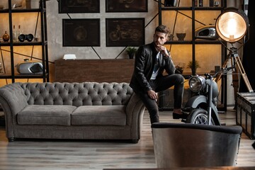 Handsome young man in leather jacket looking away while sitting on the stool