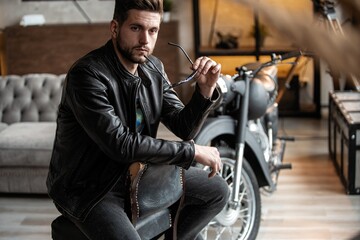 Obraz premium Handsome young man in leather jacket looking away while sitting on the stool