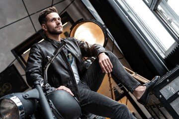 Handsome young man in leather jacket looking away while sitting on the stool