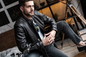Handsome young man in leather jacket looking away while sitting on the stool
