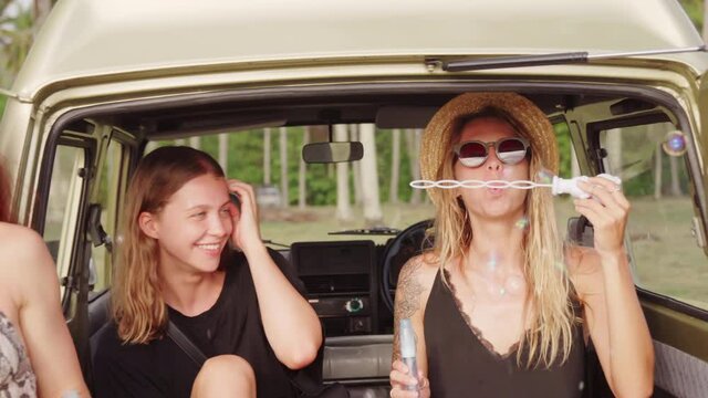 Handheld tracking of red-haired young woman and her female friends chatting and blowing bubbles together while hanging out on coast of Bali