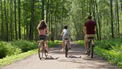 Back view follow shot of happy family of father, mother and little daughter riding bicycles together down trail in green forest - Powered by Adobe