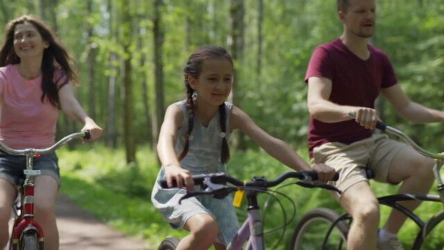 Tracking shot of happy family of mother, father and cute daughter enjoying cycling together down trail in forest