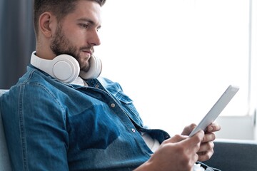 Attractive young man is using a tablet and smiling while lying on sofa at home.