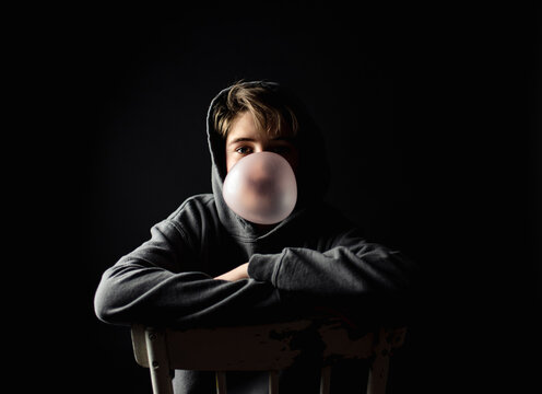 Fototapeta Teen boy in hoodie sitting on a chair in dark room blowing a bubble.