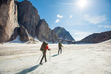 Two backpackers hiking on glacier below steep mountains.