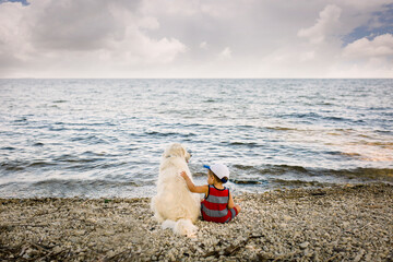 Toddler boy sitting with golden retriever dog on beach looking at lake