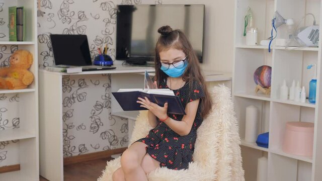 A girl with glasses and a medical mask sitting in the room on a chair and looking at the pictures in the photo album.