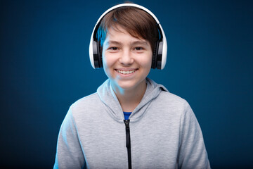Portrait of smiling young boy in hoodie. Blue background. Youngster expresses happiness. Teenager in blue headphones listening music.