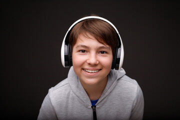 Portrait of smiling young boy in hoodie. Black background. Youngster expresses happiness. Teenager in blue headphones listening music.