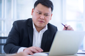 Asian businessman wore a black suit and sat in the office and looked at the laptop with stress and...