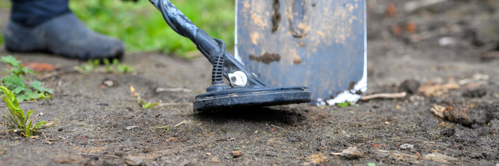 coil electronic metal detector device for search a treasure and the foots of a man on ground background and a shovel next to it. banner