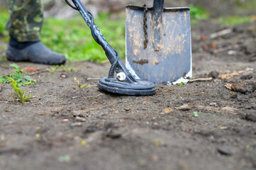 coil electronic metal detector device for search a treasure and the foots of a man on ground background and a shovel next to it