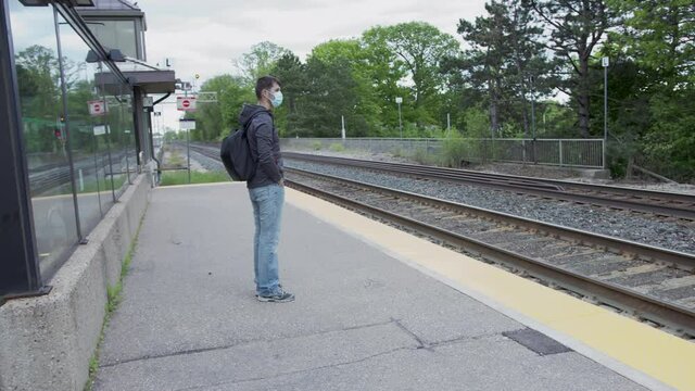 High quality 4k video of a young man who is waiting for a traing on a railway station. GO train station in Canada. Young handsome guy standing on a station and holding his cellphone wearing face mask