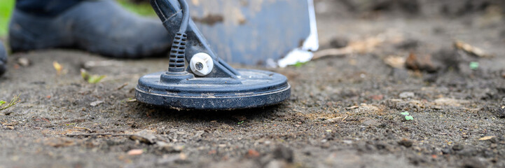 coil electronic metal detector device for search a treasure and the foots of a man on ground background and a shovel next to it. banner