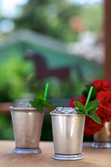 Mint Julep drinks in traditional silver cups, vertical, with red roses and horse silhouette in background.