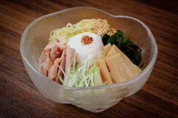 summer style cold ramen noodle with vegetables, roasted pork and poached egg in a glass bowl on...