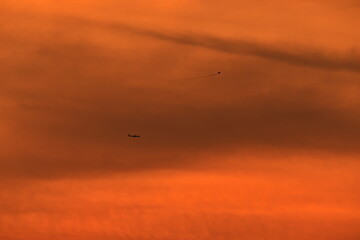 Obraz na płótnie Canvas silhouettes of an airplane and a kite, which appear to be on the same plane, flying under an orange sky during dusk.