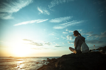 Young woman sitting on the rocky ocean coast during sunset.