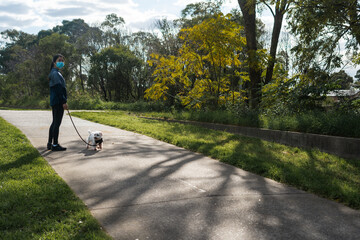 Obraz premium Girl in surgical mask in casual urban athletic wear walking with pet dog on a leash at a nature park on a footpath stylized to highlight mask during covid-19 pandemic social distancing isolation