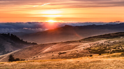 Landscape in Santa Cruz mountains, with sun rays illuminating hills covered in dry grass; People visible on the hiking trail, watching the sunset; San Francisco Bay Area, California