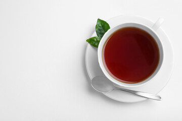 Cup of aromatic black tea, spoon and green leaves on white background, top view