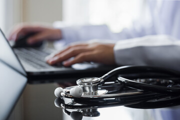 Female doctor in white coat with stethoscope working on laptop computer, using wireless mouse on the desk in medical room at clinic or hospital. Electronic health record system (EHRs) concept.