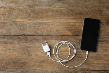 Smartphone and charging cable with adapter on wooden table, flat lay. Space for text