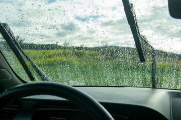 Rain drops on a car window with wipers and steering wheel looking out at the plains and the sky - obrazy, fototapety, plakaty