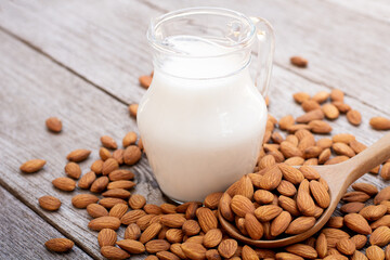 Almond milk in glass pitcher and almond nuts in wooden spoon on wood table background.