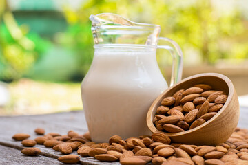 Almond milk in glass pitcher and almond nuts in wooden bowl on wood table with green nature blurred background.