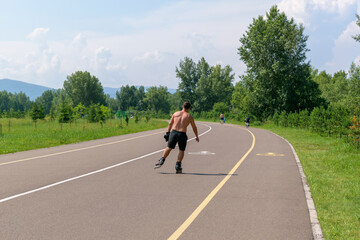 A man in shorts is rollerblading in a park. Asphalt path, green trees and grass. View from the back, muscular physique. Concept of a healthy lifestyle, sports, summer vacations.