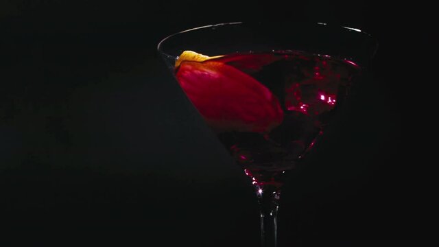 Slow motion shot of a lemon slice dropping into a  delicious cocktail glass with red liqueur infront of a black background.