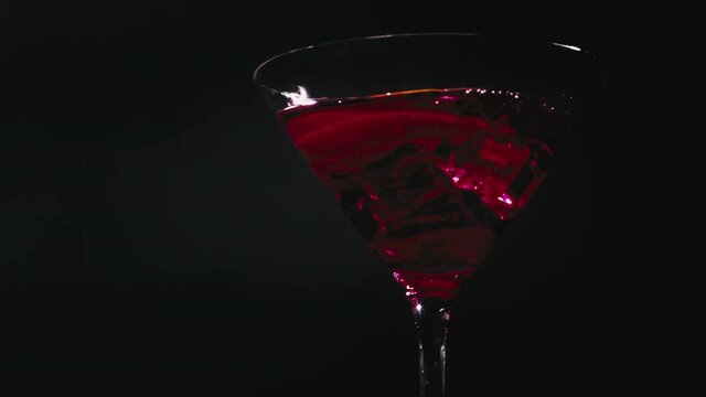 Slow motion shot of a lemon slice dropping into a delicious cocktail glass with red liqueur infront of a black background.