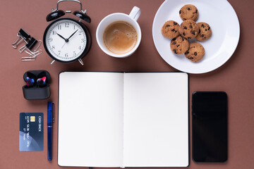 Top view - Office desk workplace with coffee cup, cookies, smartphone, credit card ,and book over brown background.