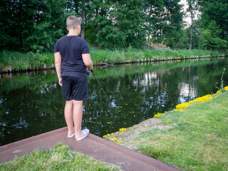 boy stands at a canal and fishes with his fishing rod