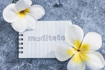 mental health concept, notepad with text Meditate next to tropical flowers on grey concrete surface