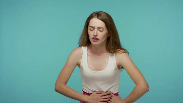 Painful cramp in stomach. Sick unhealthy girl clasping belly and suffering from severe abdominal pain, periods spasm, irritable bowel syndrome of diet. indoor studio shot isolated on blue background