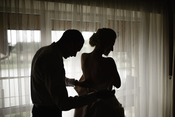 Gorgeous, blonde bride in a white luxurious dress getting ready for the wedding. Morning preparations. A woman puts on a dress. The groom helps the bride.