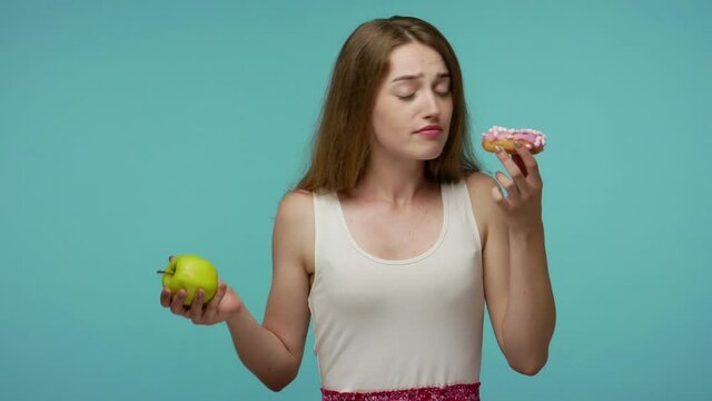 Slim beautiful girl smelling donut and biting apple, making choice between delicious fruit vs sweet sugary doughnut, vitamin nutrition concept, healthy diet. studio shot isolated on blue background